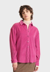 Camisa de pana rosa con frente abotonado, cuello extendido y textura acanalada. Mangas largas con detalles de botones en los puños.