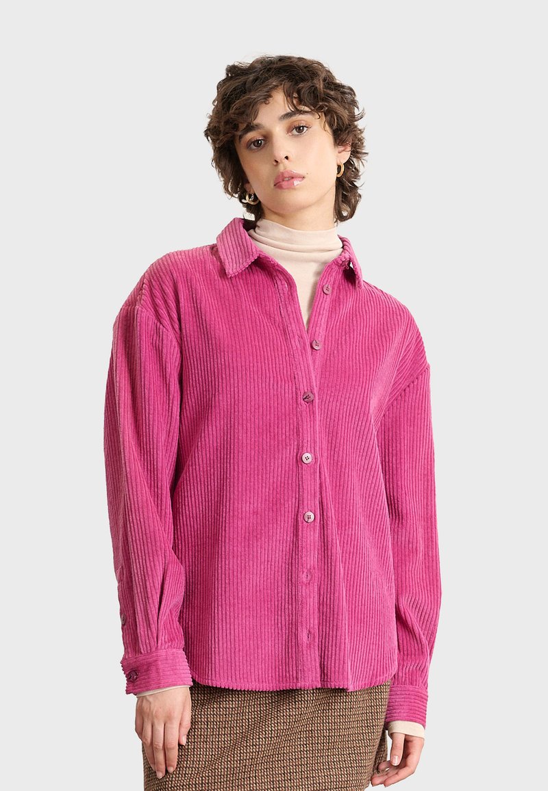 Camisa de pana rosa con frente abotonado, cuello extendido y textura acanalada. Mangas largas con detalles de botones en los puños.