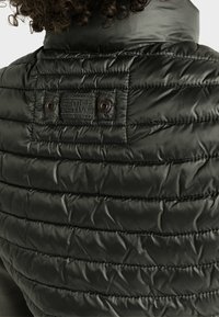 Vadderad mörkgrön väst med en kviltad struktur, utrustad med ett logomärke och knapplösningar på baksidan av kragen. Lätt och isolerad design.