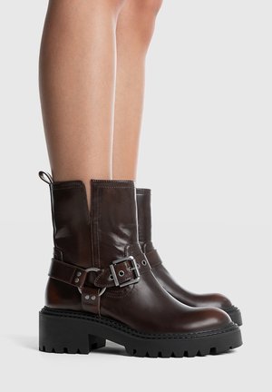 Bottes basses en cuir marron avec une semelle noire épaisse, ornées d'une boucle en métal et de détails de couture. Le dessus présente un design fendu.
