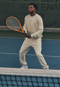 White athletic sweatshirt and matching joggers, holding an orange tennis racket, positioned on a blue tennis court with a net in foreground.