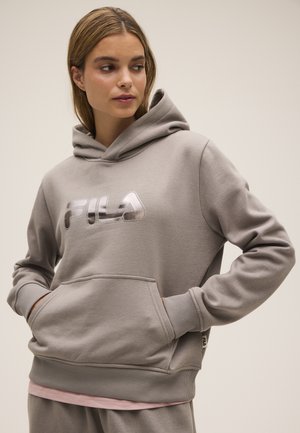 Joven mujer con sudadera con capucha taupe de FILA con logo metálico, de pie con las manos en el bolsillo delantero, mirando hacia un lado.