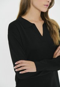 Svart långärmad topp med kragdesign och v-ringning. Materialet verkar ha textur, med långa, figurnära ärmar och en avslappnad passform runt torso.