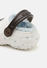 White slipper with a brown strap, featuring a soft blue fur lining and a textured sole decorated with small blue snowflakes.