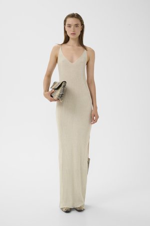 Kvinde i en beige ærmeløs lang kjole, der holder en clutch med slangeprint, iført matchende sko med slangeprint, stående mod en hvid baggrund.