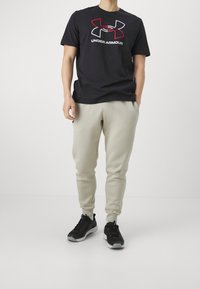 Osoba v černém tričku Under Armour, světle béžových teplácích a černých teniskách stojící před čistým bílým pozadím.