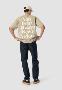 Mężczyzna w beżowej czapce, w dużym, jasnobeżowym T-shircie z napisem "Nie zawsze potrzebujesz planu" z tyłu, w ciemnych dżinsach i czarnych trampkach z gumową podeszwą.