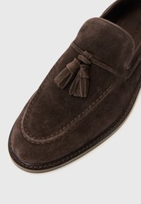 Mocassin en suede marron avec un dessus cousu, une semelle en cuir et des glands décoratifs. Texture lisse avec un design à bout arrondi.