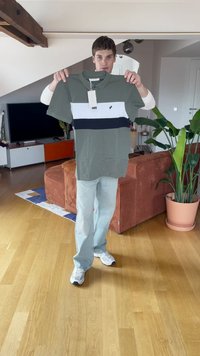 Vihreä poolopaita, jossa on valkoisia ja laivastonsinisiä vaaka-juovia, lyhyet hihat ja siihen on kiinnitetty etiketti. Malli pitää sitä kädessään huoneessa, jossa on kasveja.