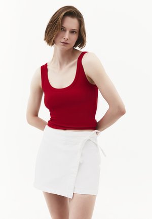 Giovane donna con capelli corti castani, che indossa un top rosso senza maniche e una gonna bianca a portafoglio, in piedi con le mani dietro la schiena.
