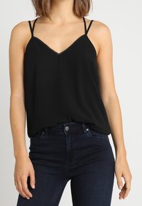 Svart ärmlös topp med tunna axelband, V-ringning och spetsdetaljer. Mjuk tyg hänger löst och kompletterar mörka denimjeans.