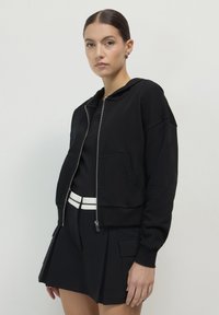 Femme portant un hoodie noir zippé, un haut noir, une jupe plissée noire avec des poches latérales et une ceinture blanche, se tenant devant un fond clair.