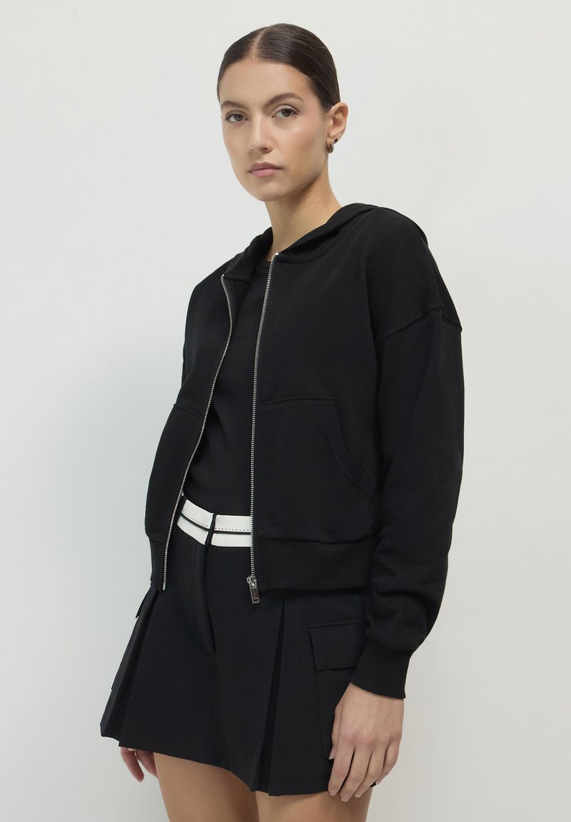 Femme portant un hoodie noir zippé, un haut noir, une jupe plissée noire avec des poches latérales et une ceinture blanche, se tenant devant un fond clair.