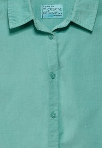 Camisa de color verde menta con cuello, confeccionada en una tela suave con textura de rayas verticales, que cuenta con un cierre de botón y una etiqueta que dice "CREATIVE GETAWAY."