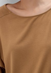 Brązowa bluzka z krótkim rękawem i szerokim dekoltem w kształcie łódki. Gładka tekstura i miękki materiał. Szwy są subtelnie widoczne wzdłuż krawędzi.