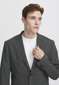 Szary żakiet z teksturowanej tkaniny, z pojedynczym zapięciem na guzik, klapami typu notch, pod którym znajduje się biały T-shirt, oraz kieszenią po lewej stronie.