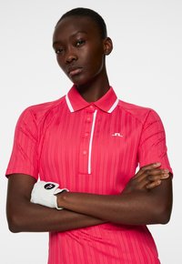 Koszulka polo w intensywnym różu z pionowymi paskami, białym kołnierzykiem i listwą. Posiada logo na piersi oraz białą rękawicę golfową na rękawie.