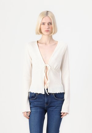 Strickoberteil mit langen Ärmeln in Creme mit tiefem V-Ausschnitt und Bindeband vorne. Hat einen fransigen Saum und einen strukturierten Stoff, kombiniert mit blauen Jeans.