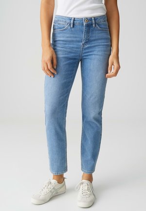 Persoon die lichtblauwe, rechte jeans draagt, witte sneakers en een witte mouwloze top, staand tegen een effen grijze achtergrond.