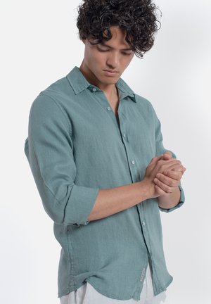 Giovane uomo con capelli ricci che indossa una camicia verde acqua abbottonata, guarda verso il basso con le mani giunte davanti a uno sfondo bianco semplice.