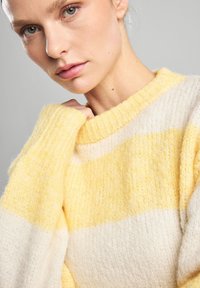 Raidallinen keltainen ja kerma väri. Pehmeä tekstuuri, ribattu kaulus ja hihansuut. Lähikuva mallin kasvoista, käsi kauluksen lähellä.