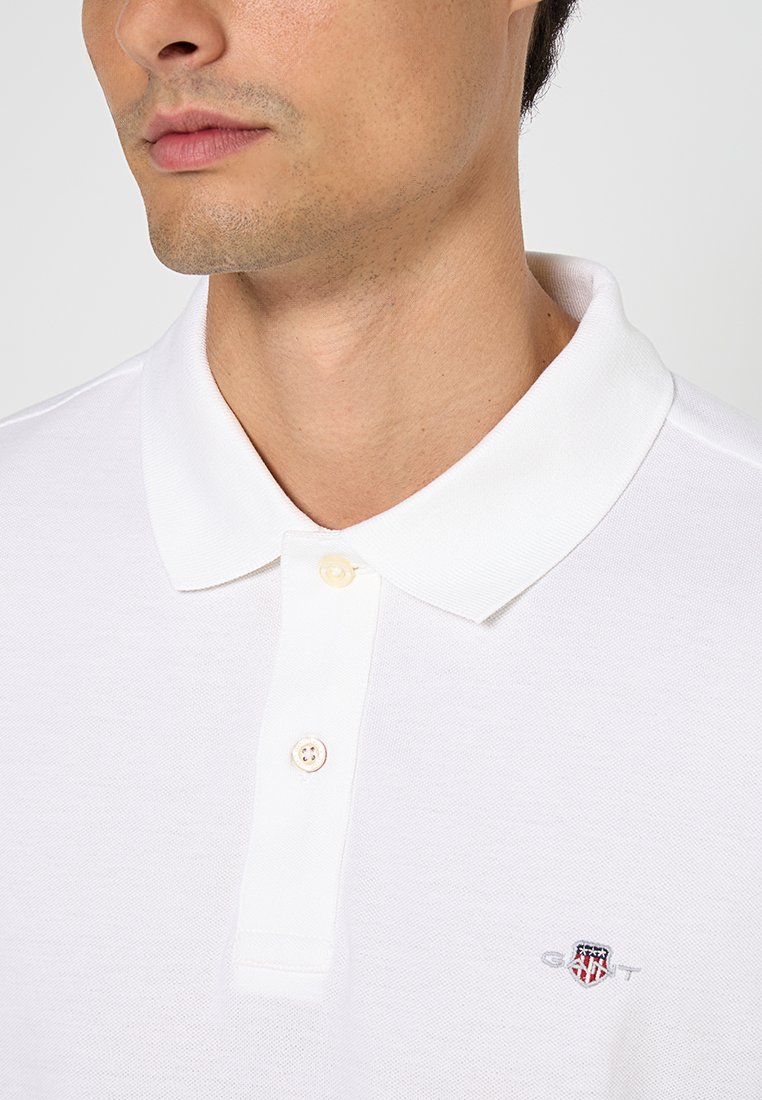 Weißes Baumwollpolohemd mit geripptem Kragen und zwei Knöpfen, ausgestattet mit einem bestickten Logo auf der linken Brust. Strukturierter Stoff, schlichtes Design.