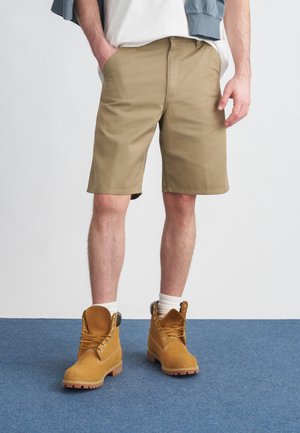 Pantalones cortos caqui hechos de tela ligera, con bolsillos laterales. Combinados con botas marrones hasta el tobillo, con cordones con detalles amarillos y suelas texturizadas.