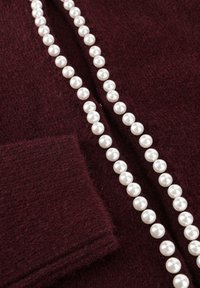 Een streng witte parels rustend op burgundy gestructureerde stof met een zichtbare manchet rand.