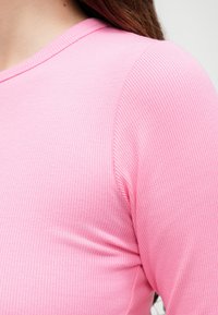 Roza rebrasta majica s mekim teksturom, okruglim izrezom i prilagođenim rukavima, naglašavajući glatku i rastezljivu tkaninu.