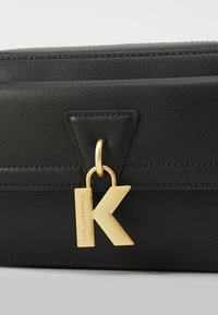 Svart lærveske med en strukturert design, med stikking og en gullfarget metalllogoformet som bokstaven "K."