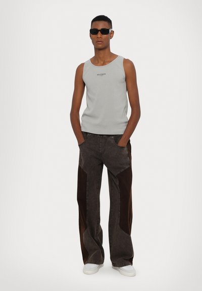 Canotta grigia a coste con la scritta "SILENCE" stampata in nero, abbinata a jeans a gamba larga marrone scuro con pannelli laterali marrone chiaro e sneakers bianche.
