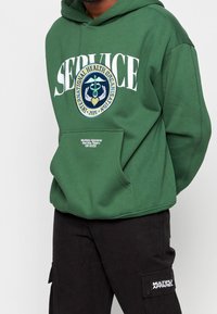 Felpa verde con testo bianco "SERVICE" e un emblema che rappresenta un caduceo, tasca frontale e tessuto morbido. Pantaloni neri con tasche cargo.