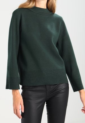 Femme portant un pull vert foncé à manches longues avec un col montant et un pantalon en cuir noir sur un fond uni.