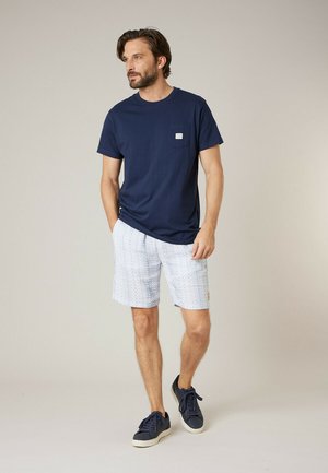 Homme portant un t-shirt bleu marine avec une poche, un short clair à motif, et des baskets bleu marine, debout devant un fond uni.