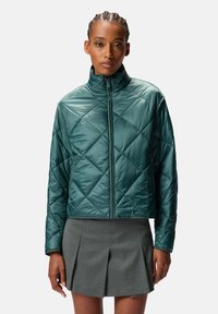 Giacca imbottita verde con collo alto, cerniera frontale e finitura lucida, indossata sopra una gonna grigia plissettata.