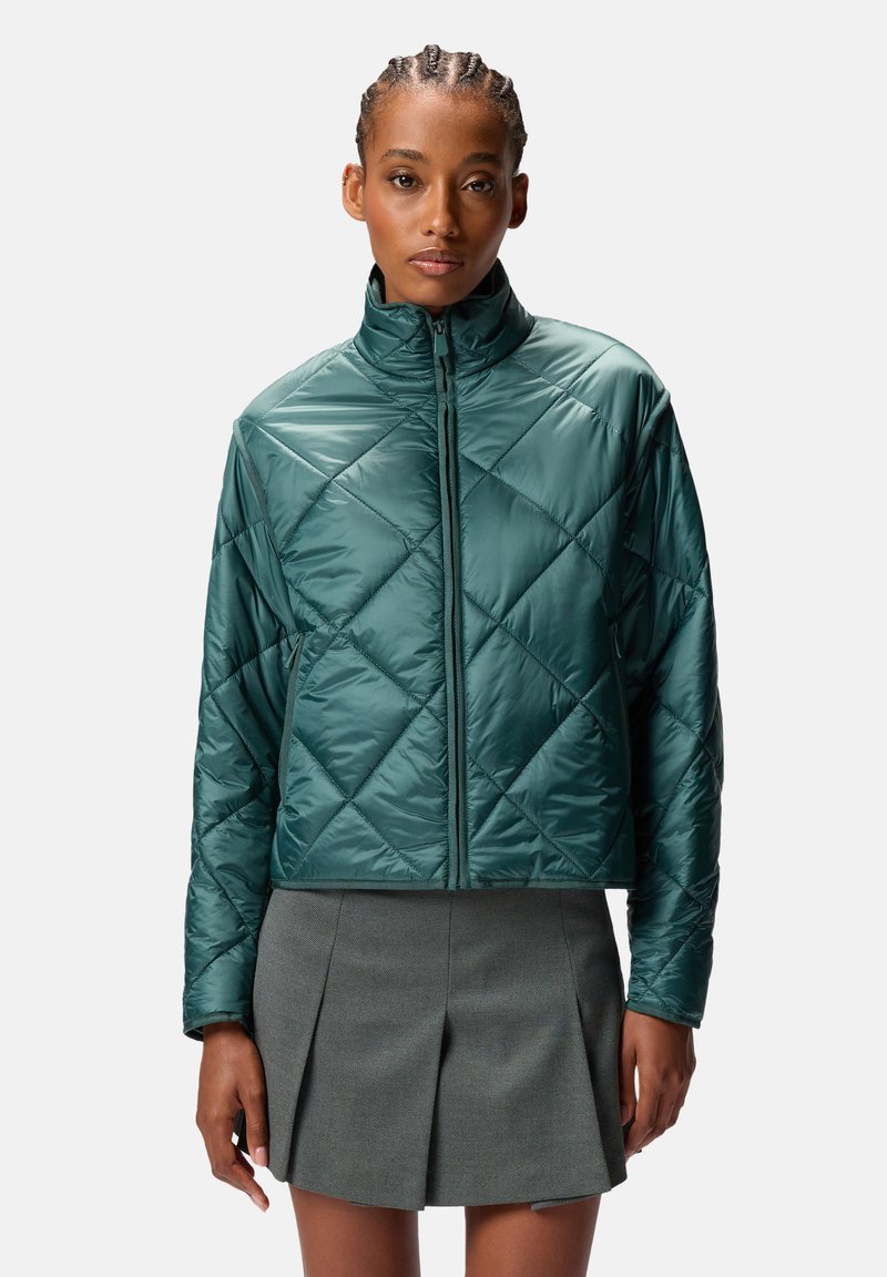 Giacca imbottita verde con collo alto, cerniera frontale e finitura lucida, indossata sopra una gonna grigia plissettata.