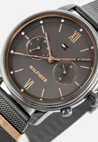 Montre avec bracelet en maille noire, cadran gris foncé, accents dorés, index en chiffres romains et trois sous-cadrans, arborant le logo "HILFIGER."