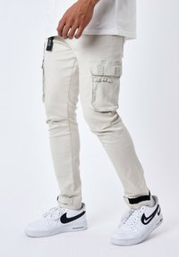 Pantalones cargo beige de corte ajustado, con bolsillos laterales y detalle de lazo. Combinados con zapatillas blancas y una camiseta blanca básica.