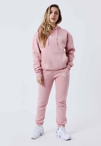 Felpa con cappuccio rosa, dotata di tasca frontale e testo ricamato bianco, abbinata a pantaloni della tuta coordinati con polsini elasticizzati e dettagli con logo.