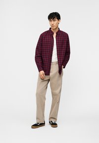 Νέος άνδρας που στέκεται, φορώντας πουκάμισο με καρό σε μπορντό και ναυτικό μπλε πάνω από λευκή φανέλα, μπεζ παντελόνι και μαύρα αθλητικά παπούτσια με λευκές κορδόνια.