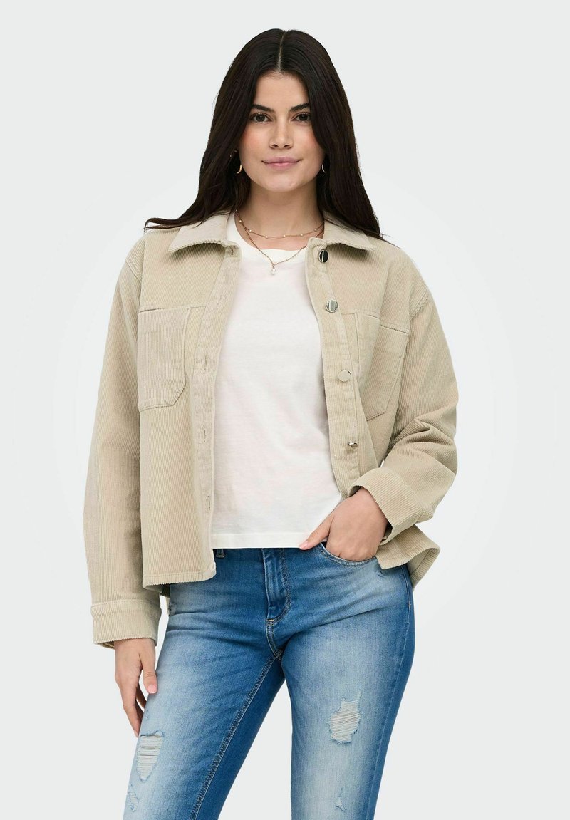 Femme aux longs cheveux foncés portant une veste beige en velours côtelé boutonnée sur une chemise blanche et un jean bleu déchiré, debout avec une main dans la poche.