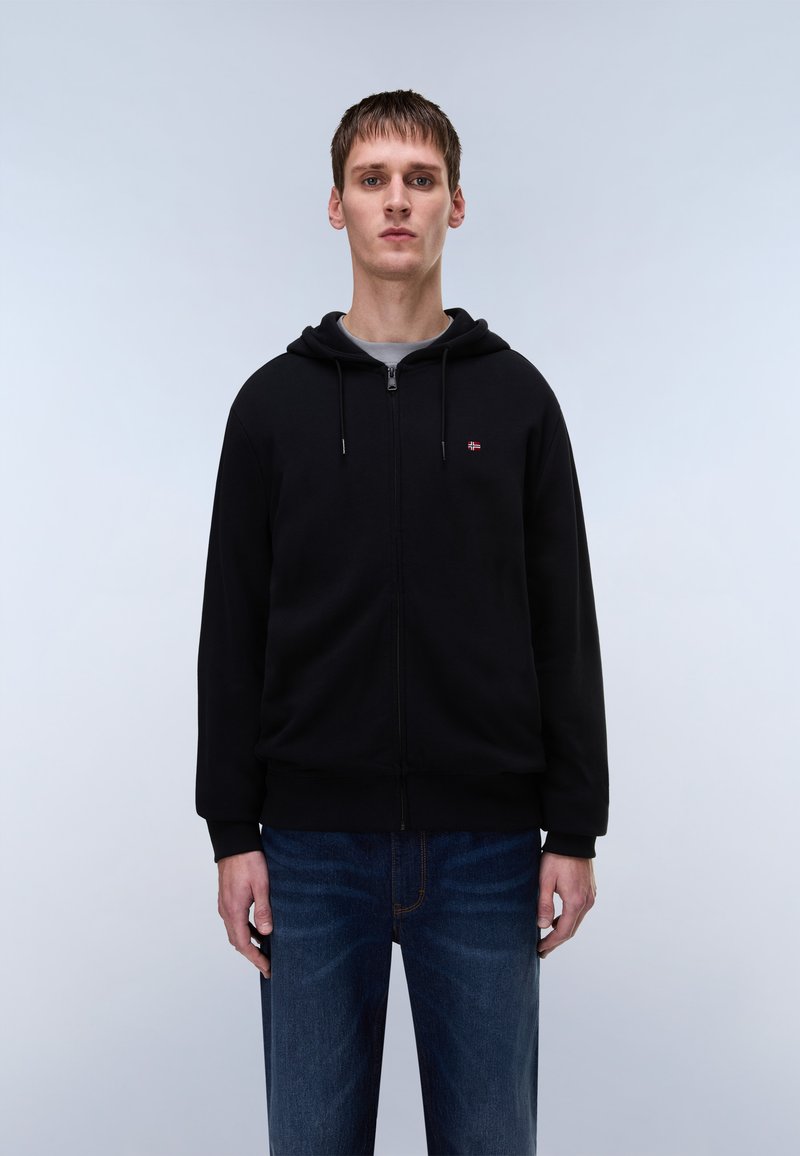 Must zipuga kapuutsiga pusa esitasku ja reguleeritava paelaga kapuutsiga, mille juurde on kombineeritud tumedat sinist teksapükse, millel on peen tekstuur ja õmblused.