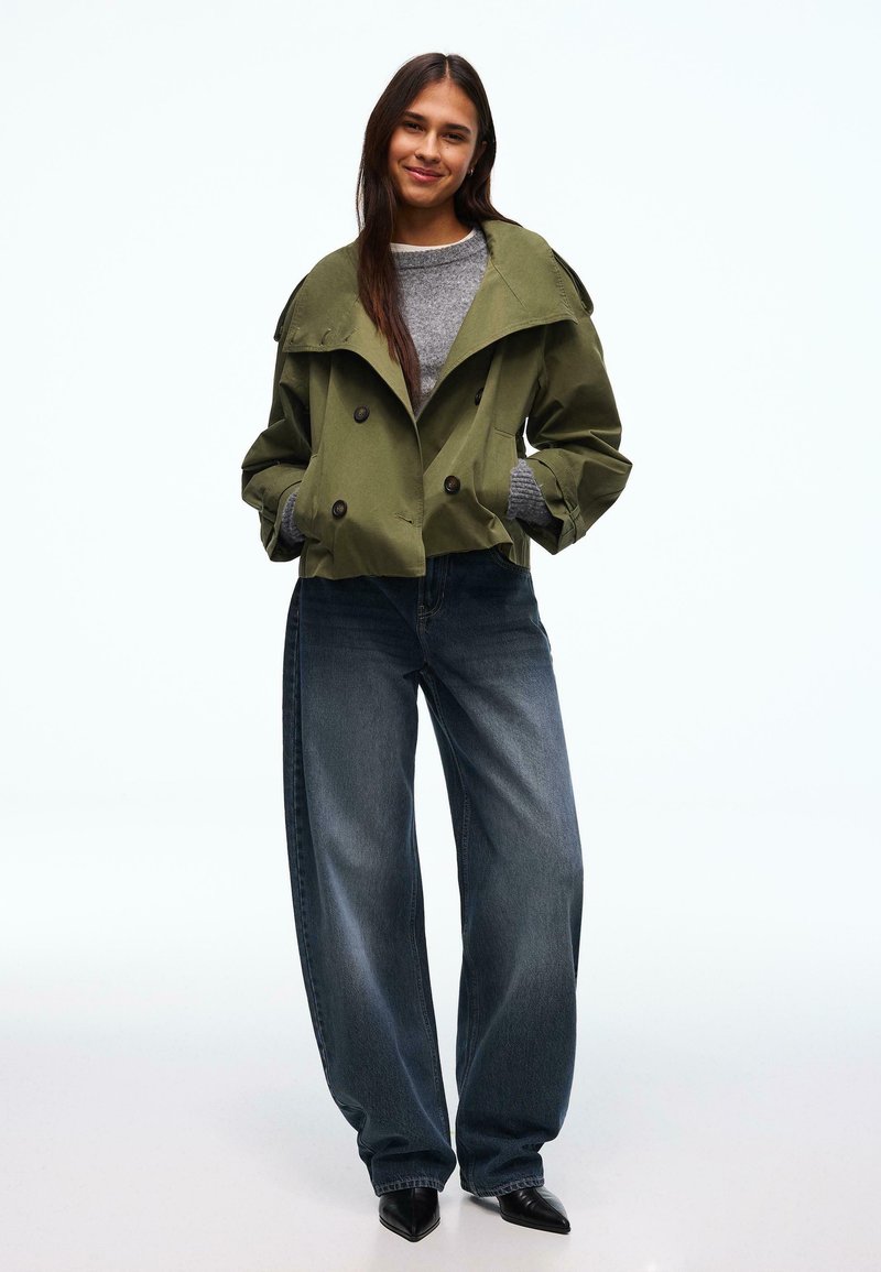 Trench coat verde oliva doppiopetto con ampio colletto, indossato sopra un maglione grigio. Abbinato a jeans di denim scuro a gamba larga e scarpe nere con punta appuntita.