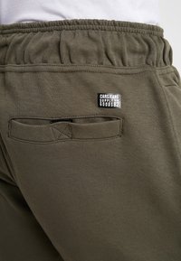 Olijfgroene joggers van een gestructureerde stof, met een elastische tailleband en een klein zwart label met de tekst "CARSJEANS SUPPLY GOODS82" nabij een achterzak.