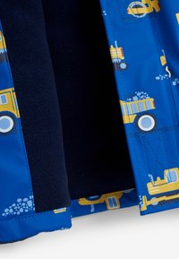 Jachetă albastră cu modele de camioane galbene. Are căptușeală din fleece și exterior matlasat, cu cusături decorative și interior contrastant în nuanță de navy.