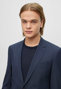 Giacca da uomo blu navy con revers a punta e taschino singolo, abbinata a un maglione in maglia scura. Tessuto liscio, vestibilità tailor-made.