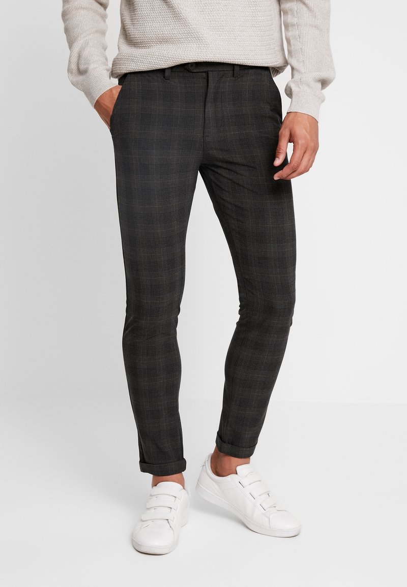 Homme portant un pantalon slim gris foncé à carreaux, un pull beige et des baskets blanches, debout avec une main dans la poche.