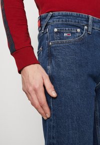 Mörkblå jeans i denim med regular fit, synlig sömnad, framfickor och en logopatch på baksidan av midjan. Hand vila på benet.