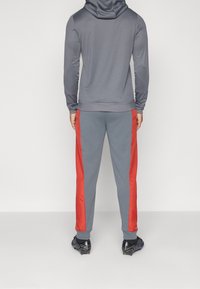 Persona de espaldas con una sudadera gris y pantalones de chándal gris a juego con rayas rojas a los lados, de pie sobre un suelo blanco.