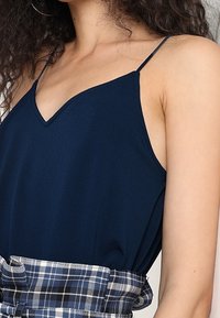 Sininen ilman hihasia toppi, jossa on V-pääntie ja kevyt kangas, yhdistettynä sinivalkoiseen ruudulliseen vyöhön, tuoden lisätekstuuria.